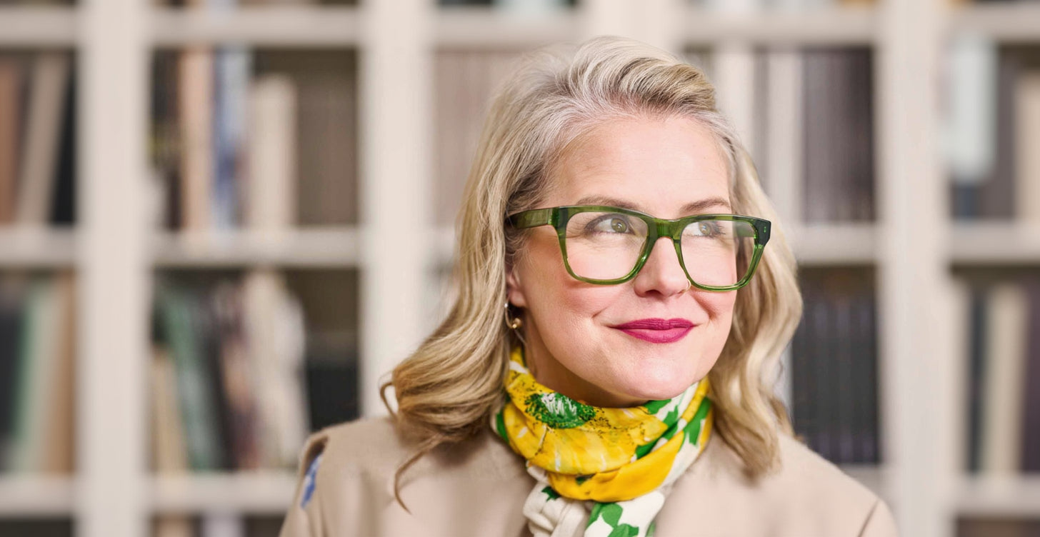 Woman wearing green glasses and a colorful scarf in front of a blurred bookshelf.
