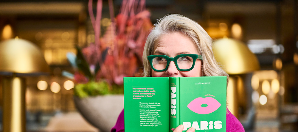 Person holding a green book titled 'Paris' in a blurred indoor setting